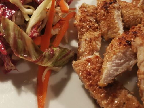 Strisce di pollo impanato con semi di sesamo servite accanto a un'insalata di radicchio e carote a julienne.