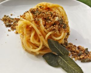 Un nido di spaghetti conditi con una cremosa salsa alla zucca, guarniti con granella di noci, semi di girasole e foglie di salvia fritta su un piatto bianco.