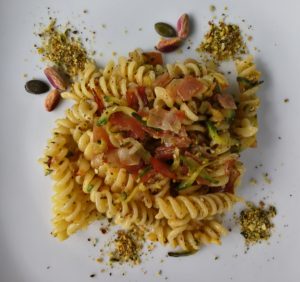 Un piatto di fusilli conditi con zucchine a julienne, speck a listarelle, granella di pistacchi e semi di zucca su un piatto bianco.