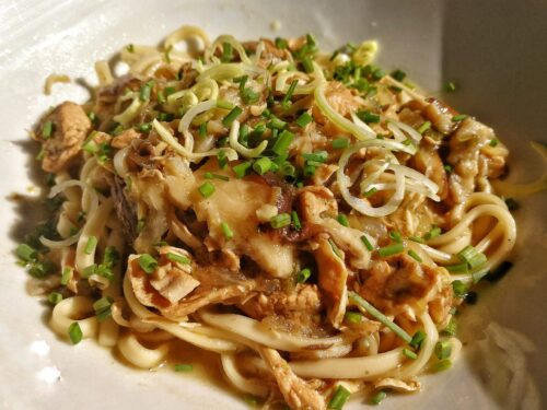 Primo piano di un piatto di noodles saltati con pezzi di pollo, funghi shiitake e guarnizione di erba cipollina fresca.