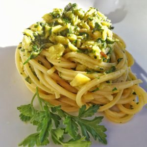 Un piatto di spaghetti conditi con pezzetti di stoccafisso e bietole verdi, guarnito con un rametto di prezzemolo fresco su un piatto bianco.