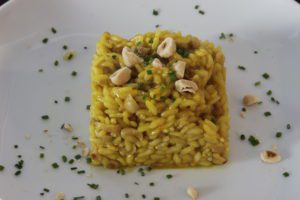 Un piatto di risotto allo zafferano di colore giallo intenso, guarnito con nocciole intere e erba cipollina tritata, servito su un piatto bianco.