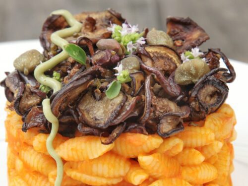 Un primo piano di gnocchetti sardi conditi con pomodorini datterini arancioni, sormontati da melanzane saltate, capperi e una decorazione di mousse verde.