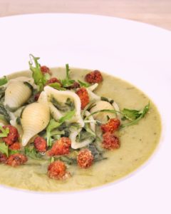 Un piatto di pasta formato abissine servito su una vellutata verde di piselli e porri, guarnito con cicoria fresca e croccanti pezzetti di 'nduja fritta.