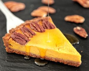 Una fetta di tart di zucca guarnita con noci pecan su cui viene versato dello sciroppo, servita su un piatto scuro.