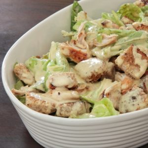 Una ciotola bianca riempita con insalata Cesare condita, contenente lattuga fresca, pezzi di pollo grigliato e crostini di pane croccanti.