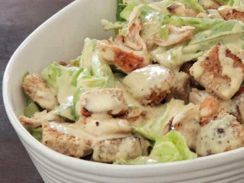 Una ciotola bianca riempita con insalata Cesare condita, contenente lattuga fresca, pezzi di pollo grigliato e crostini di pane croccanti.