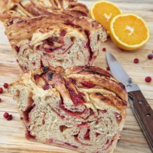 Una fetta di babka all'arancia e mirtilli rossi su un tagliere di legno accanto a un coltello e fette di arancia fresca.