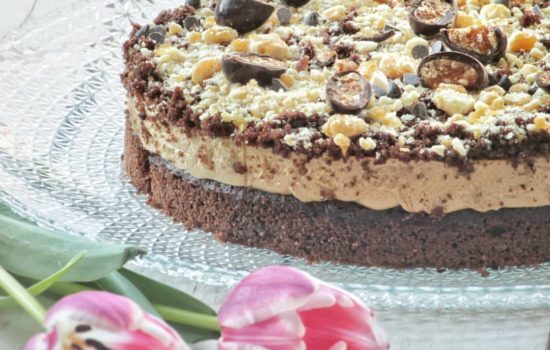 Una torta al cioccolato e caffè guarnita con granella e cioccolatini su un piatto di vetro, con due tulipani rosa in primo piano.