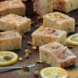 Quadrotti di brownies vegani al limone glassati e guarniti con granella di pistacchi, disposti su una superficie scura insieme a fette di limone.