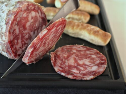 Una mano taglia una fetta di salame stagionato su un tagliere nero, accanto a piccoli pezzi di pane.
