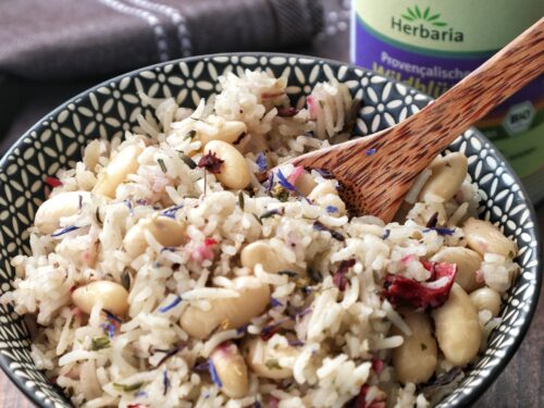 Una ciotola decorata contenente riso basmati con fagioli cannellini e fiori edibili, servito con un cucchiaio di legno accanto a una confezione di spezie Herbaria.