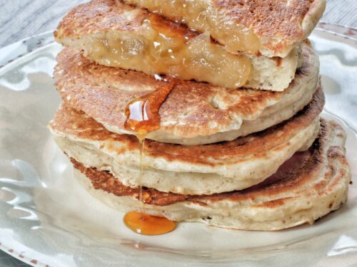 Una pila di pancake vegani ripieni di mele su un piatto, guarniti con sciroppo d'acero che cola.