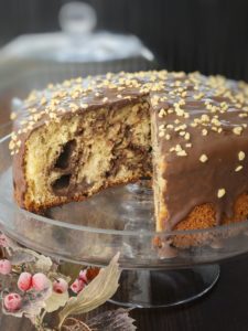 Una torta rotonda con glassa al cioccolato e granella di nocciole su un'alzatina di vetro, con una fetta mancante che rivela l'interno soffice e variegato.