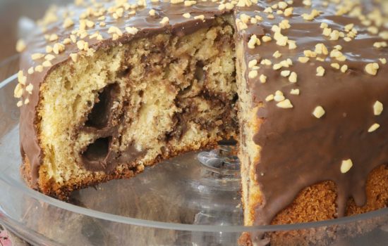 Una torta soffice con glassa al cioccolato e granella di nocciole, servita su un'alzatina di vetro con una fetta tagliata.