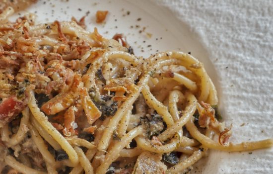 Un piatto di spaghetti conditi con una crema di carciofi, cavolo riccio viola e guanciale croccante su un piatto bianco.