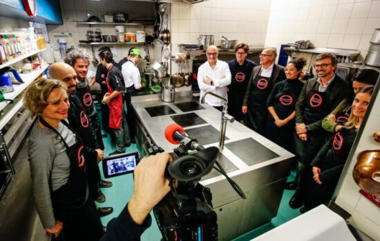 Un gruppo di persone indossa grembiuli neri con il logo ArchiChef in una cucina professionale, ripresi da una videocamera.