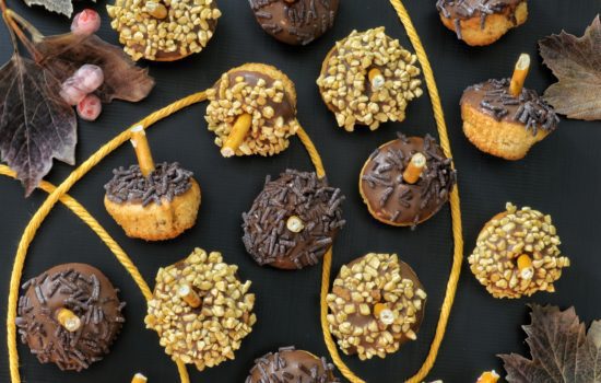 Piccoli dolcetti a forma di ghianda decorati con glassa al cioccolato, granella di nocciole e codette, disposti su un piano scuro con foglie autunnali.
