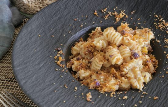Un piatto di pasta radiatori condita con cavolfiore e una granella croccante, servito su un piatto nero opaco.