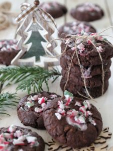 Una pila di biscotti vegani al doppio cioccolato guarniti con granella di bastoncini di zucchero alla menta, disposti su un tavolo con decorazioni natalizie.