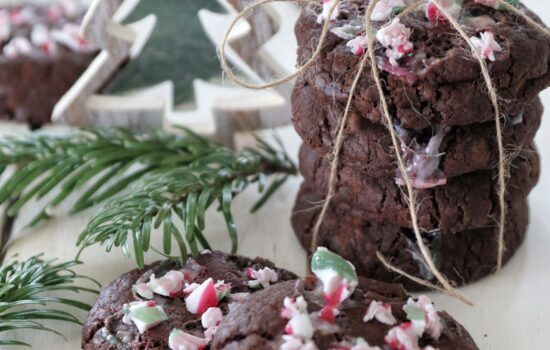 Una pila di biscotti vegani al doppio cioccolato decorati con granella di bastoncini di zucchero alla menta, disposti su un tavolo con decorazioni natalizie.