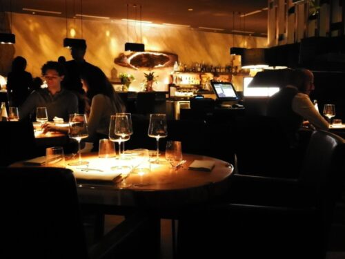Interno di un ristorante con tavoli apparecchiati e clienti seduti in un ambiente con illuminazione soffusa.
