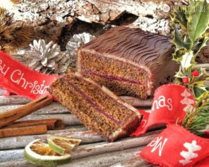 Una fetta di tronchetto natalizio farcito con confettura e ricoperto di marzapane e cioccolato fondente, posizionato su una base decorata con cannella, agrifoglio e nastri rossi.