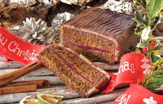 Una fetta di tronchetto natalizio farcito con confettura e ricoperto di marzapane e cioccolato fondente, decorato con nastri rossi e agrifoglio.
