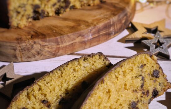 Due fette di Pan d'Arancio vegano con gocce di cioccolato su un piano di legno, con il resto del dolce sullo sfondo.