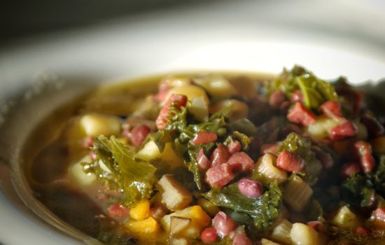 Un piatto fondo contenente una zuppa calda con cubetti di prosciutto, fagioli azuki, carote gialle, carciofi e cavolo riccio.