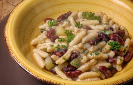 Un piatto di gnocchetti sardi conditi con baccalà, carciofi e fagioli rossi, servito in una ciotola di ceramica gialla su un tavolo di legno.