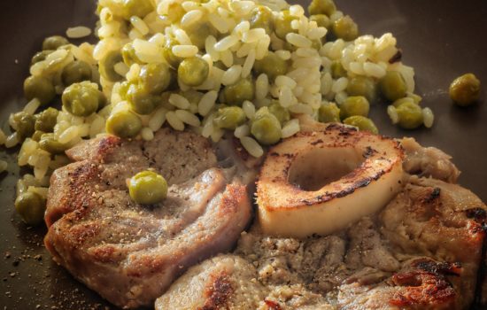 Un piatto di ossobuco fumante servito con un contorno di riso e piselli su un piatto scuro.