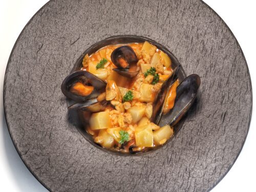 Un piatto fondo scuro contenente una zuppa di riso, patate e cozze con un sugo al pomodoro.