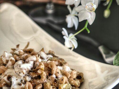 Un piatto di pasta a forma di trecce condito con carciofi e una salsa cremosa al guanciale, servito in un piatto bianco moderno accanto a un'orchidea bianca.