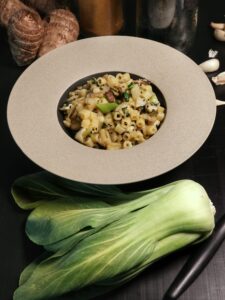 Un piatto di pasta corta saltata con Pak Choi, funghi e semi di nigella, servito in una ciotola dal bordo ampio su un tavolo scuro.