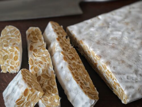 Un panetto di tempeh tagliato a fette su un tagliere di legno scuro, con un coltello da cucina sullo sfondo.