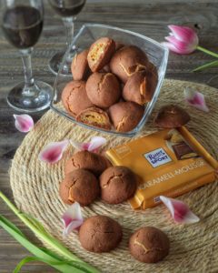 Una ciotola di vetro piena di biscotti al cioccolato accanto a una tavoletta di cioccolato Ritter Sport Karamell-Mousse su un sottopentola di paglia decorato con petali di tulipano.