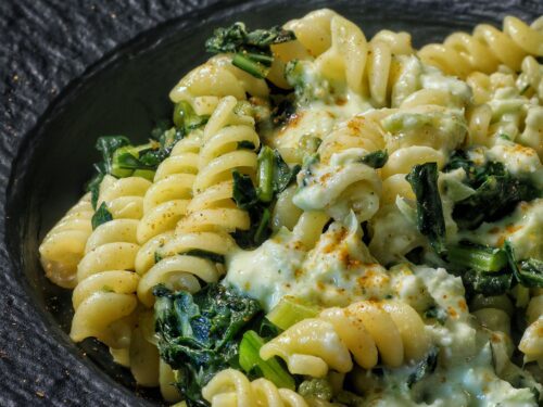 Un primo piano di fusilli conditi con foglie di cavolo rapa e una cremosa salsa al pecorino romano, serviti in un piatto scuro.