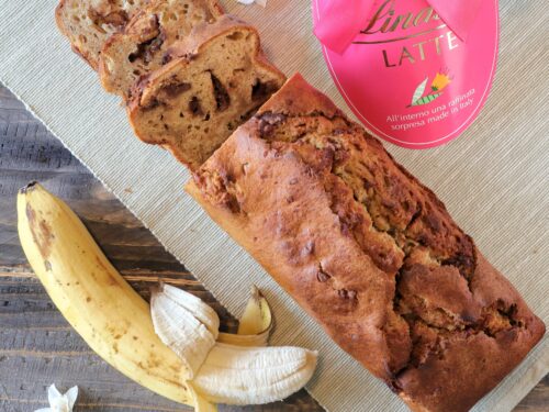 Un banana bread tagliato a fette su un tovagliolo di stoffa accanto a una banana sbucciata e una confezione regalo di cioccolato Lindt.