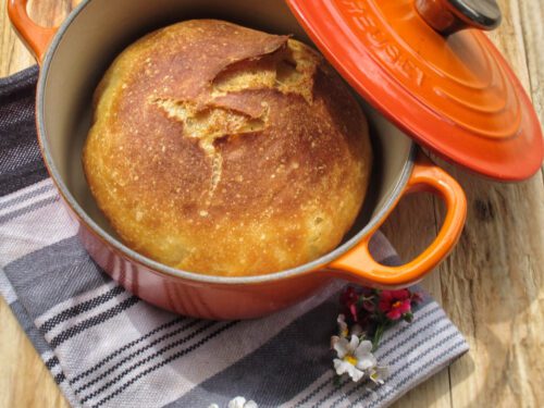 Una pagnotta di pane appena sfornata all'interno di una cocotte in ghisa arancione Le Creuset appoggiata su un canovaccio a righe.