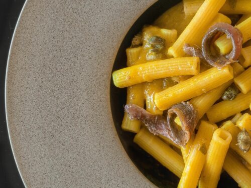 Un piatto di pasta corta condita con una salsa ai pomodorini gialli, guarnita con filetti di acciuga arrotolati e capperi.