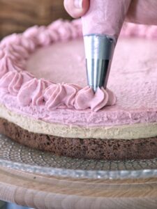 Una mano utilizza una sac à poche per decorare con ciuffi di crema rosa la superficie di una torta a strati.