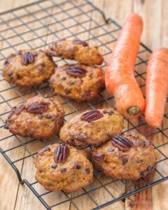Biscotti vegani alle carote arricchiti con gocce di cioccolato e noci pecan, disposti su una griglia di raffreddamento accanto a due carote fresche.