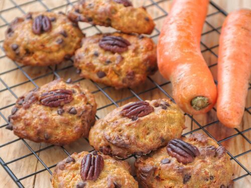 Biscotti vegani alle carote arricchiti con gocce di cioccolato e noci pecan, disposti su una griglia di raffreddamento accanto a due carote fresche.