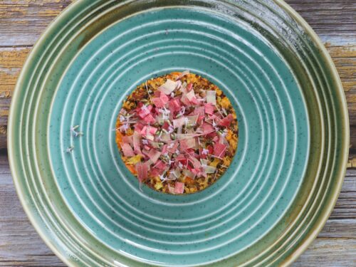 Un piatto circolare in ceramica verde acqua con una porzione di quinoa guarnita con pezzetti di rabarbaro e piccoli fiori, poggiato su un tavolo di legno rustico.