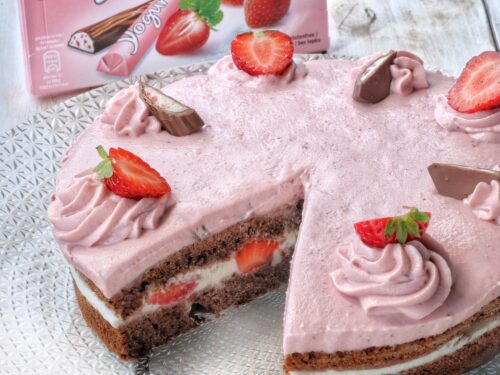 Una torta al cioccolato farcita con crema rosa e fragole fresche, decorata con ciuffi di crema e pezzi di barrette Yogurette, posizionata davanti alla confezione originale del prodotto.