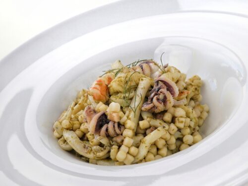 Un piatto di fregula condita con seppie, mazzancolle e pesto verde, servito in un piatto fondo bianco.