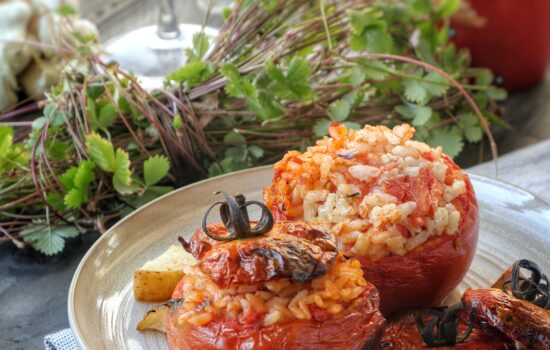 Due pomodori maturi ripieni di riso cotto al forno, serviti su un piatto di ceramica chiara con erbe aromatiche fresche sullo sfondo.
