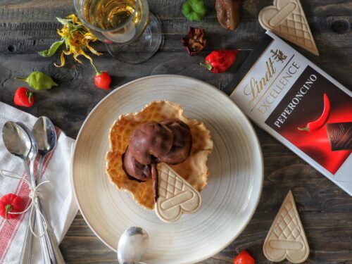 Una coppa di gelato al cioccolato servita in una cialda su un piatto, accompagnata da una tavoletta di cioccolato Lindt al peperoncino e peperoncini freschi su un tavolo di legno.
