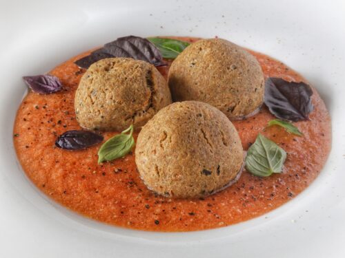 Tre polpette croccanti adagiate su una base di gazpacho rosso, guarnite con foglie di basilico verde e viola.